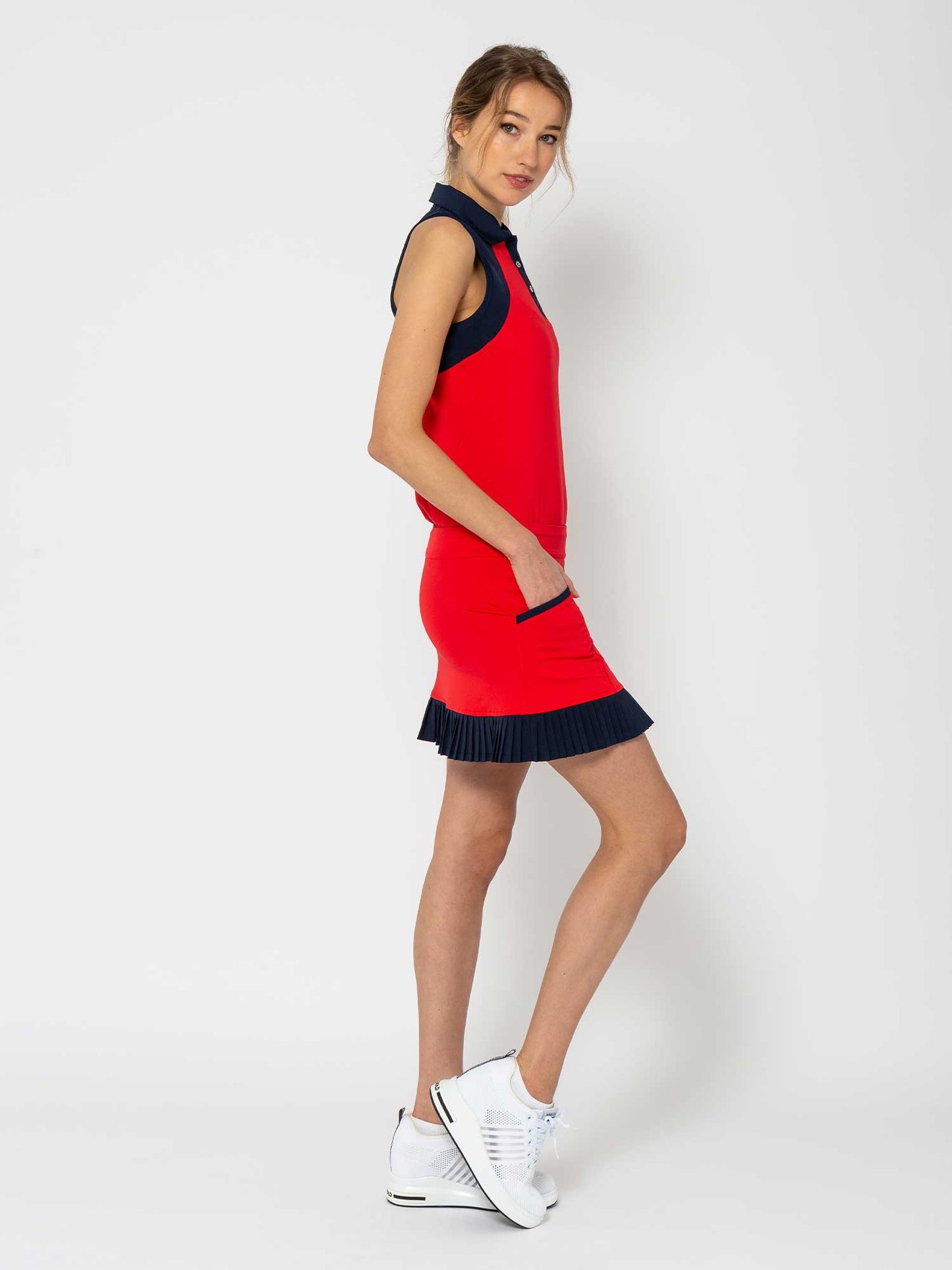 A woman stands sideways against a white background wearing the inPhorm NYC Kai 17" Pleated Skirt in Poppy/Midnight, styled like a golf skirt with UV protection. She has her hands in the pockets, wears white sneakers, and her light brown hair is loosely tied back.