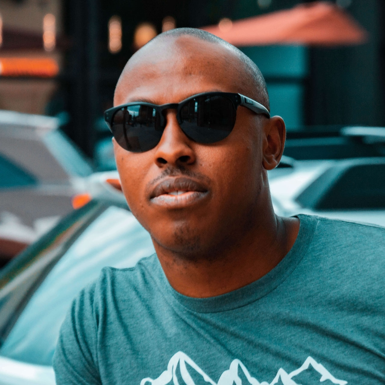 A man with a shaved head wears Epoch Eyewear's Sierra sunglasses and a green tee with a white mountain graphic outdoors. Sunlit, blurred cars and buildings in the background create an urban vibe.