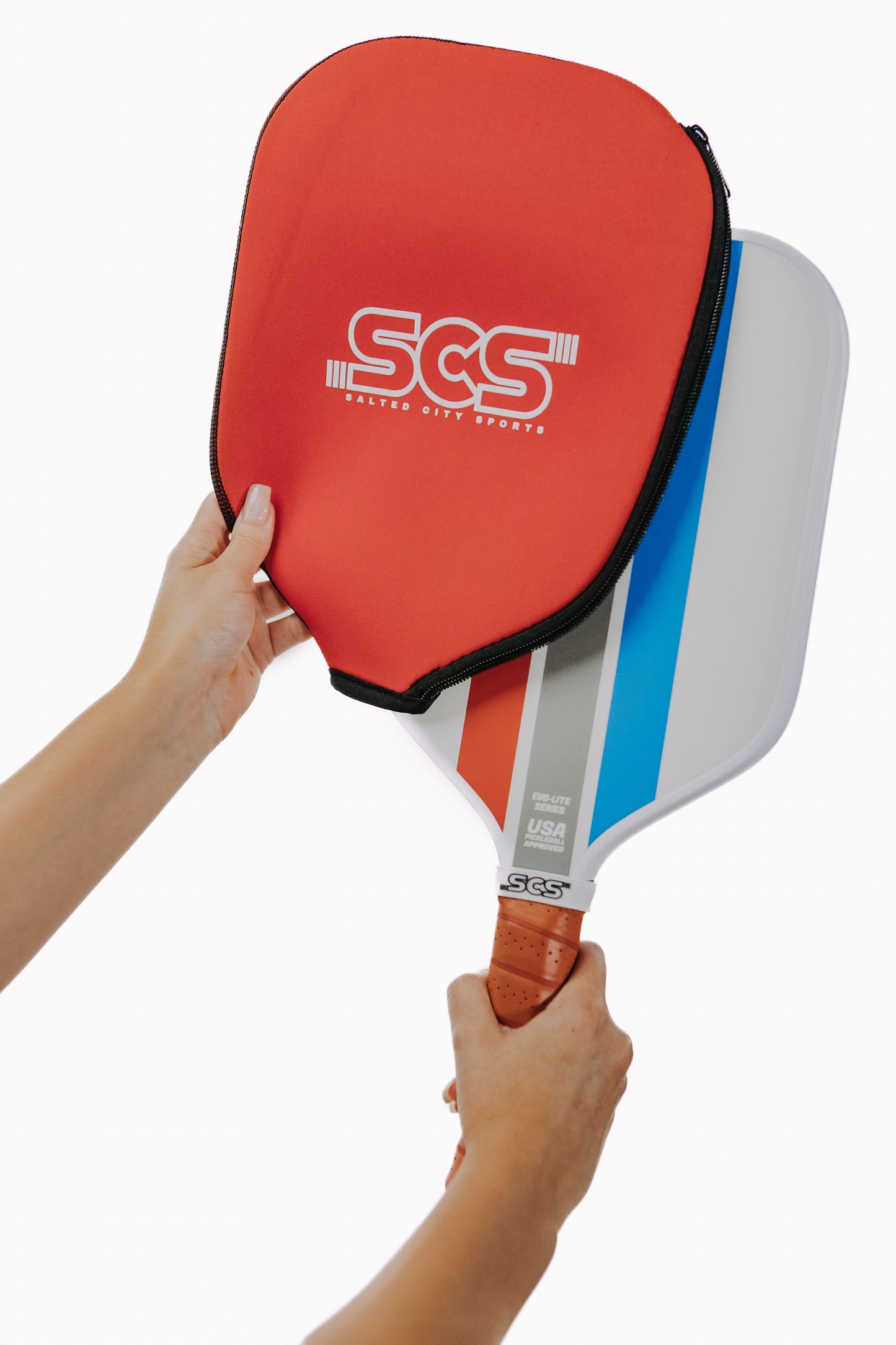 A person holds a Salted City Sports Mav | Evo-Lite Series pickleball paddle with a polypropylene core and tan grip, featuring red and blue stripes. In the other hand, they place a red Salted City Sports cover over the paddle.