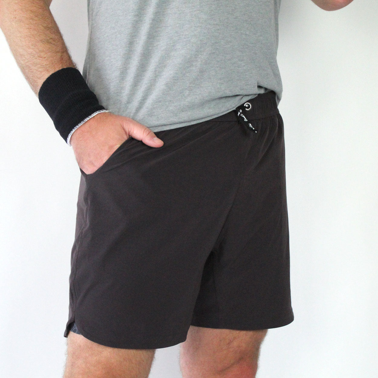 A person wears a light grey t-shirt, Good Get Pickleball Shorts, GG Edition with a compression liner, and a black wristband. They stand with one hand in their pocket against a white background; only their torso and upper legs are shown.