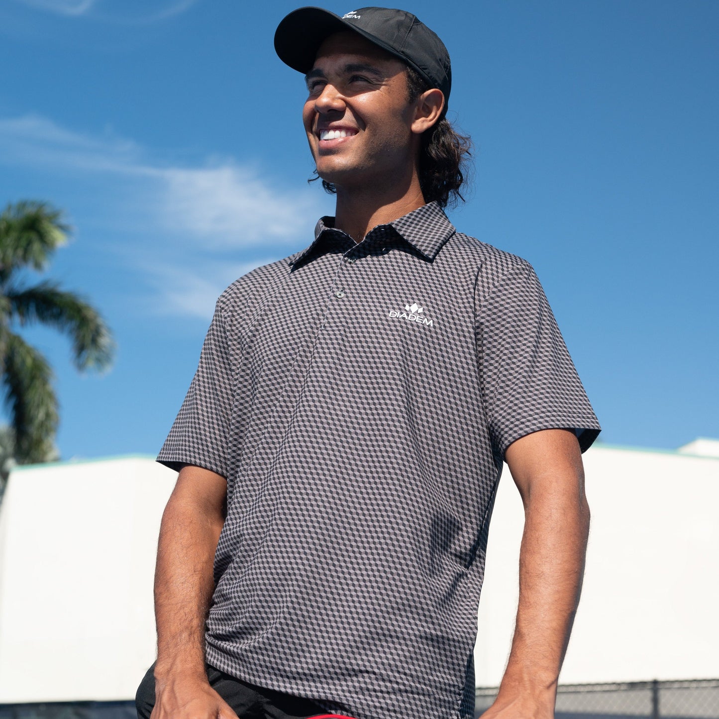 A young man in a black hat and Diadem Sports Men's Scales Competition 18K Pattern Polo smiles outdoors under a clear blue sky. A palm tree and white building hint at a warm, sunny day—perfect for moisture wicking comfort.