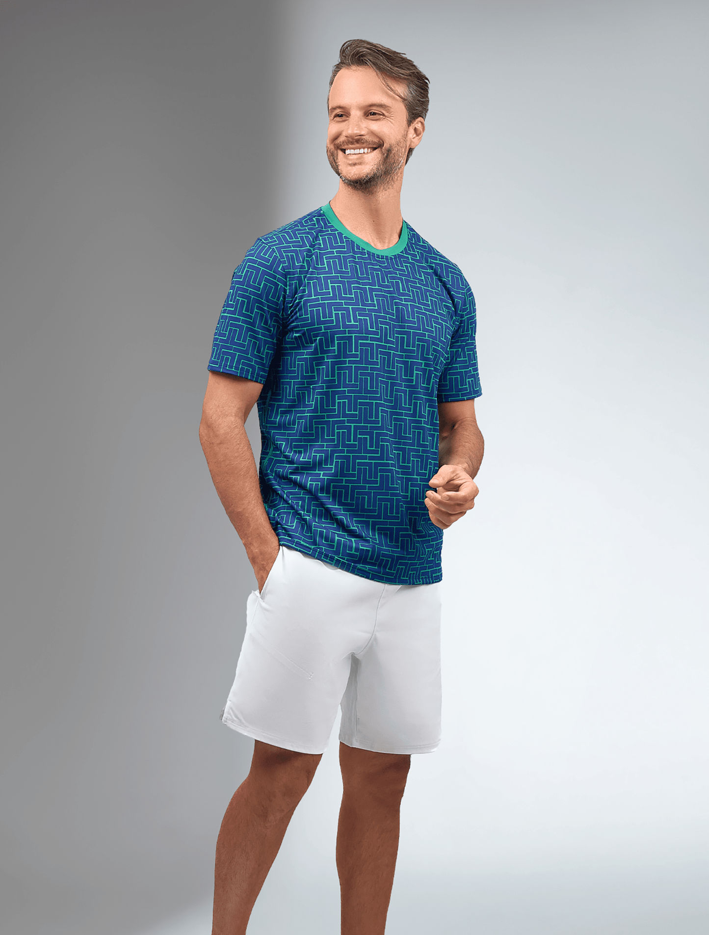 A smiling man with short brown hair stands with one hand in his pocket, wearing the inPhorm NYC Men's Short-Sleeve Crew Neck with UV protection and antimicrobial features, paired with white shorts against a gray-to-white gradient background.