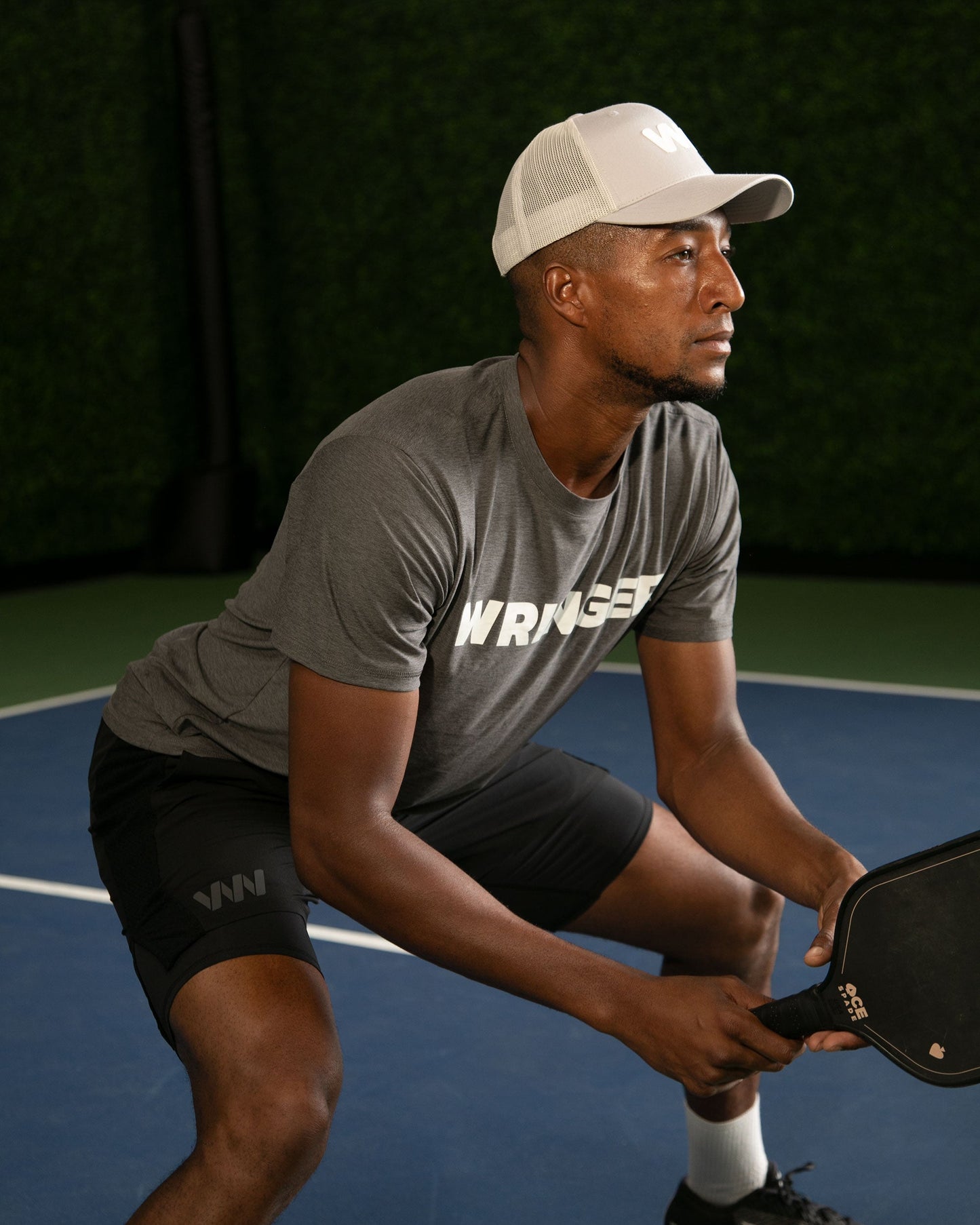 A man in a gray Wringer Wear Men's Short Sleeve WRINGER T-shirt, black shorts, and a white cap crouches on a blue pickleball court with a paddle in hand. A lush green hedge wall is visible in the background.