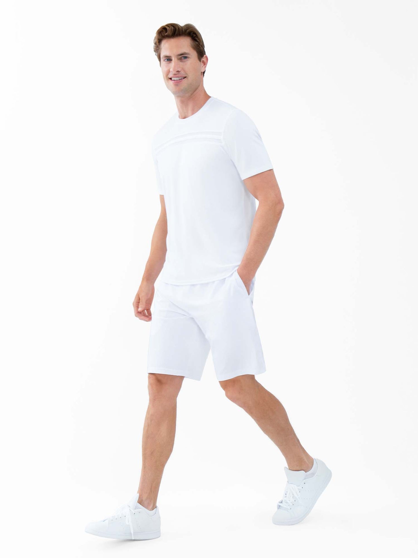 A man with light skin and short brown hair walks and smiles, wearing the inPhorm NYC Performance Men's Tournament Short Sleeve Crew Neck in white. The quick-dry, moisture-wicking shirt stands out against a clean, minimalistic white background.
