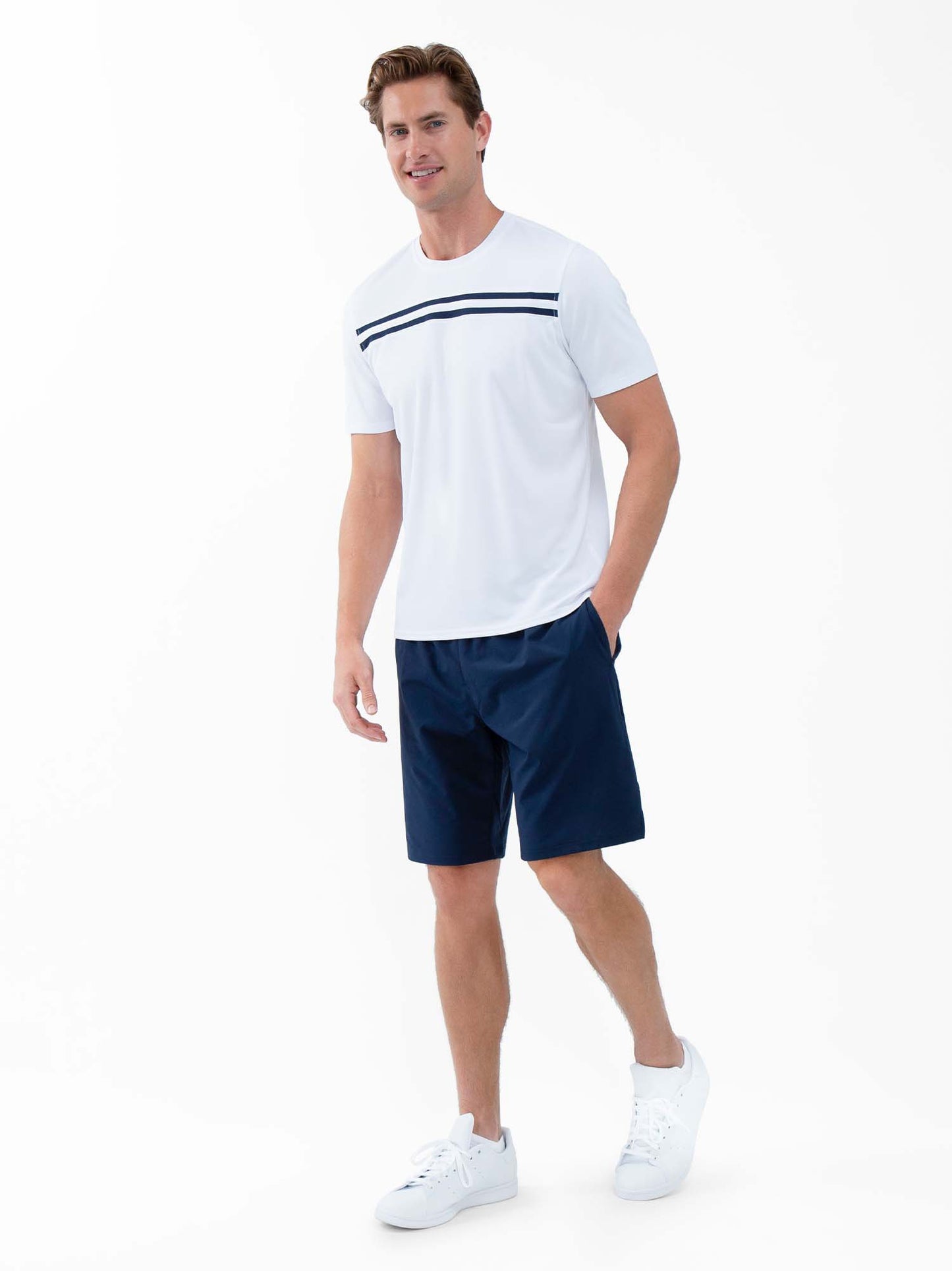 A man with short light brown hair smiles as he walks forward, wearing the inPhorm NYC Performance Men's Tournament Short Sleeve Crew Neck in White/Midnight and dark navy shorts, paired with white sneakers. One hand rests casually in his pocket.