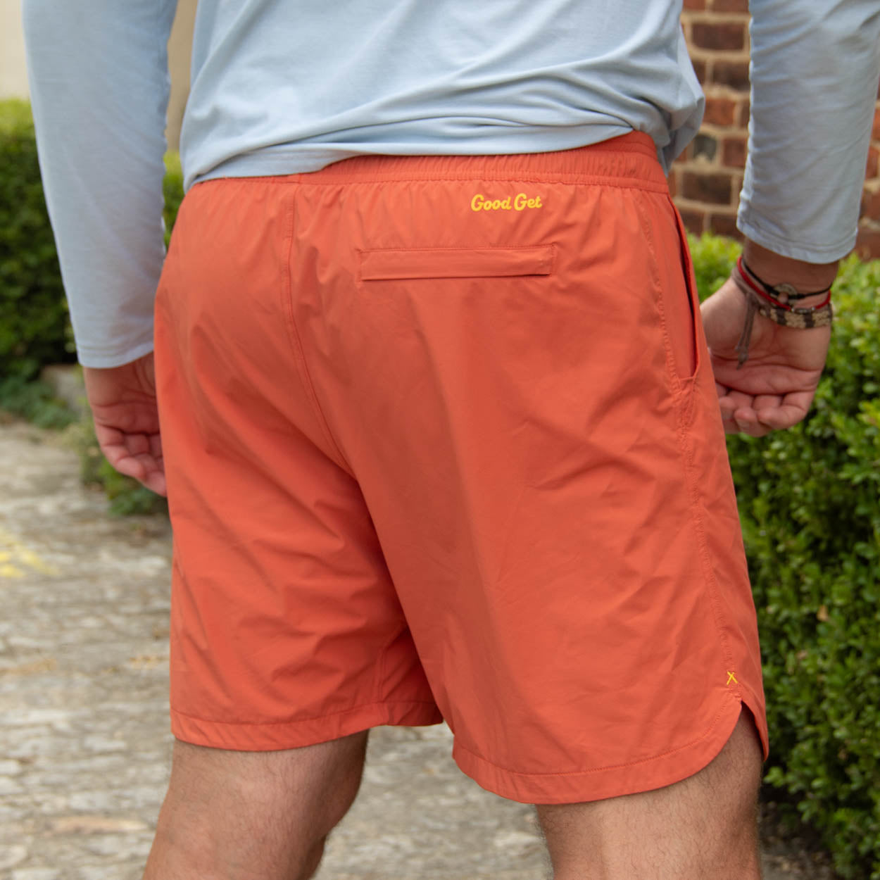 A person stands outside with their back to the camera, wearing Good Get Pickleball Shorts, Drippy Edition in orange. "Good Get" is printed in yellow above the back pocket. They pair these 6-way stretch shorts with a light blue long-sleeved shirt and bracelets.