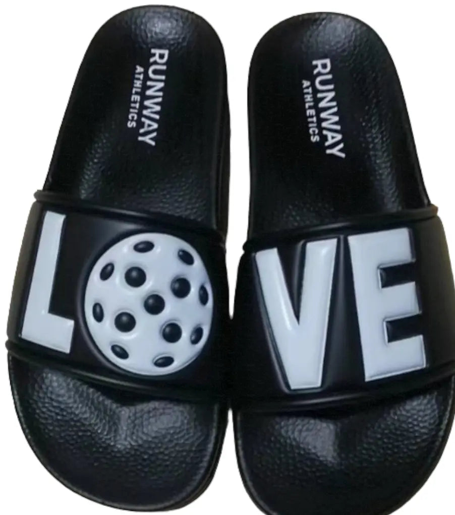 A pair of black Pickleball Slides by Runway Athletics features custom straps: the left shows "L" with a 3D white pickleball as the "O," and the right displays "V" and "E," spelling out "LOVE" in raised letters when worn together.