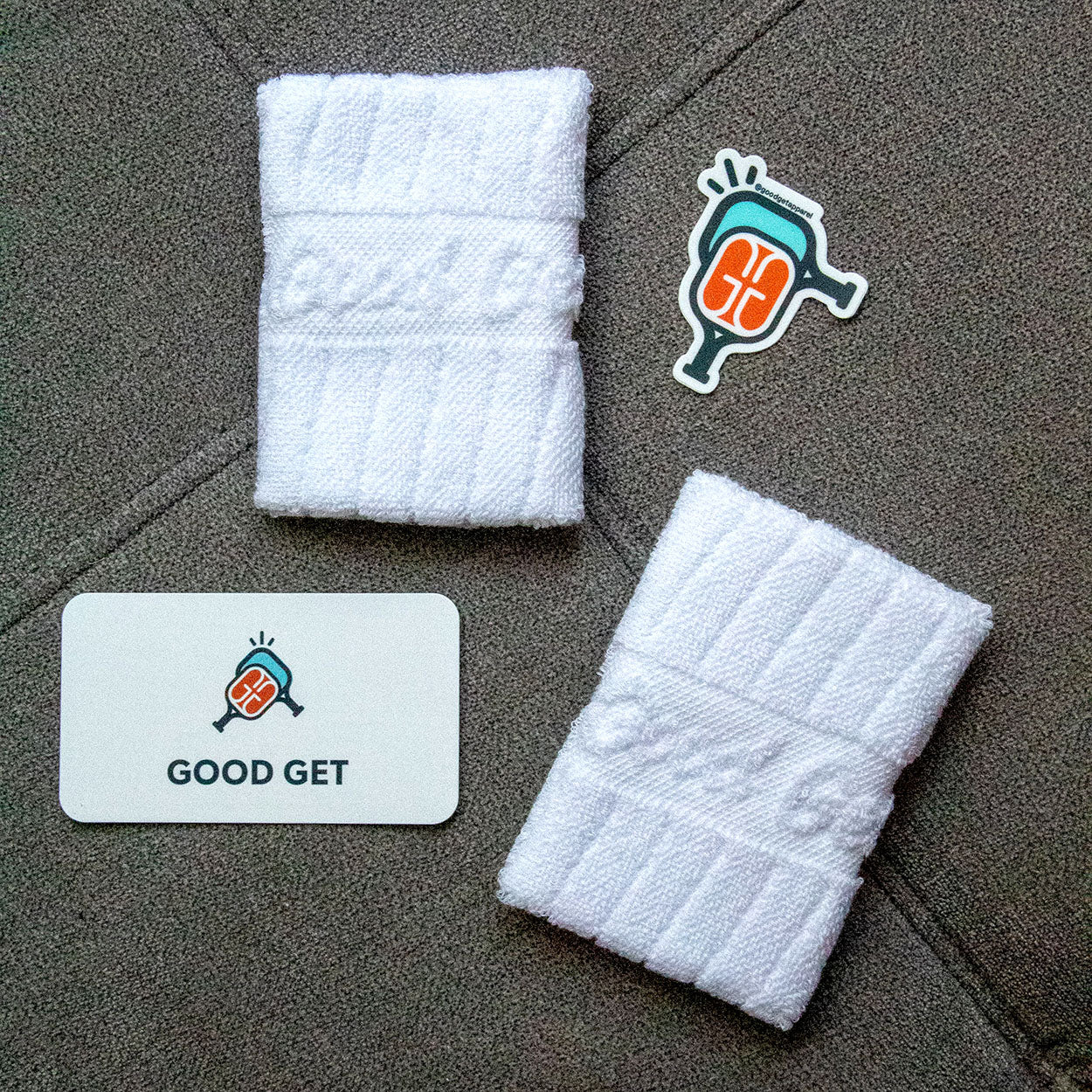 Two white Good Get Wristband of Dinking +1 terry cloth wristbands rest on a gray textured surface, with a sticker and card featuring a stylized paddle hitting a ball and the GOOD GET logo placed between them.