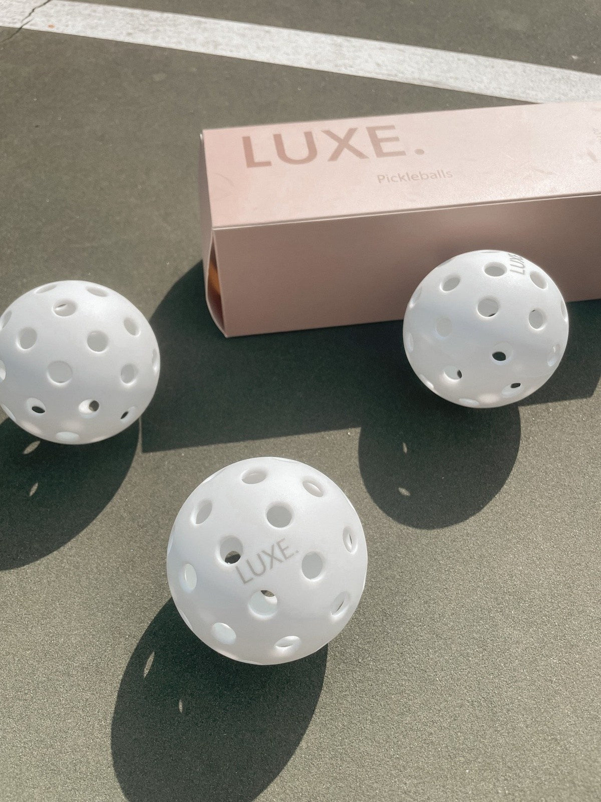 Three white LUXE Pickleballs by LUXE Pickleball sit on an outdoor court near a light pink box. Each durable ball, ideal for indoor and outdoor play, features multiple holes and a LUXE stamp, with soft sunlight casting shadows nearby.