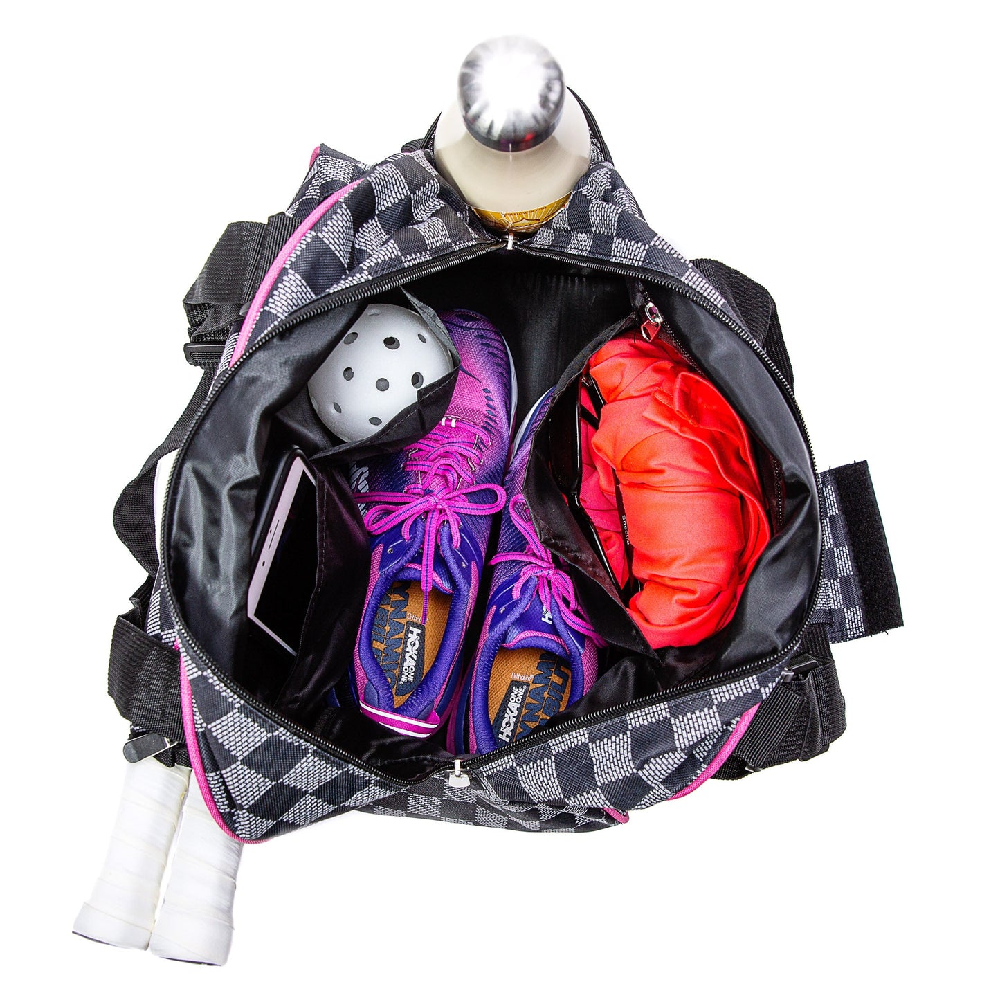 The Pik'le'Ball Sports Checkered Chic Premium Women's Pickleball Bag with padded compartments and adjustable strap, shown from above with pink trim, holds a white ball, purple sneakers, orange garment, smartphone, water bottle, and two side grips.