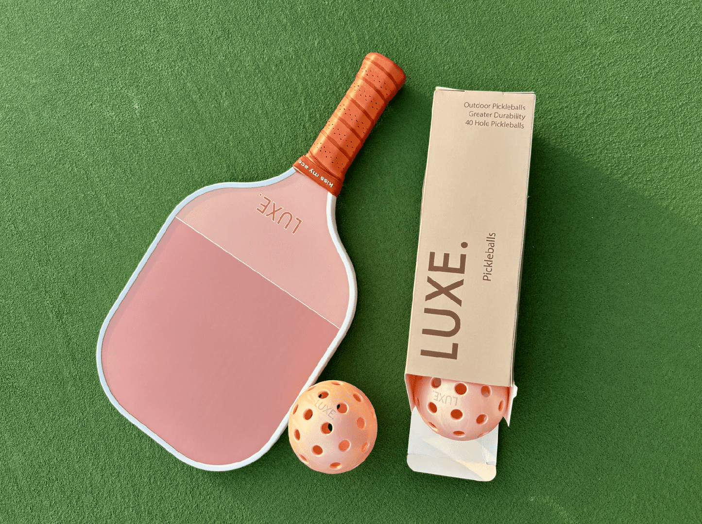 A Pink Pickleballs - Pack of 3 by LUXE Pickleball is displayed with a pink paddle featuring an orange grip on a green surface. Two orange 40-hole balls are shown, one inside and one outside the branded box.