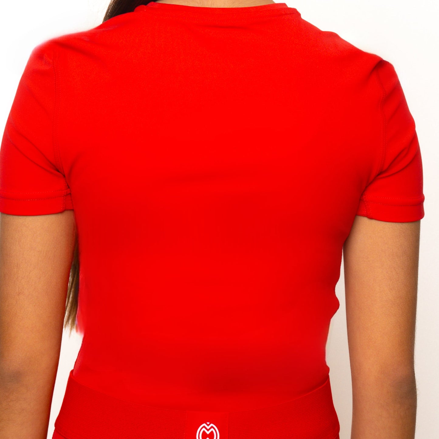 A girl with long hair is seen from behind wearing a fitted Massov ProForm® Girls' Short Sleeve Compression Shirt in red, featuring a small white logo near the bottom hem, shown against a plain white background.