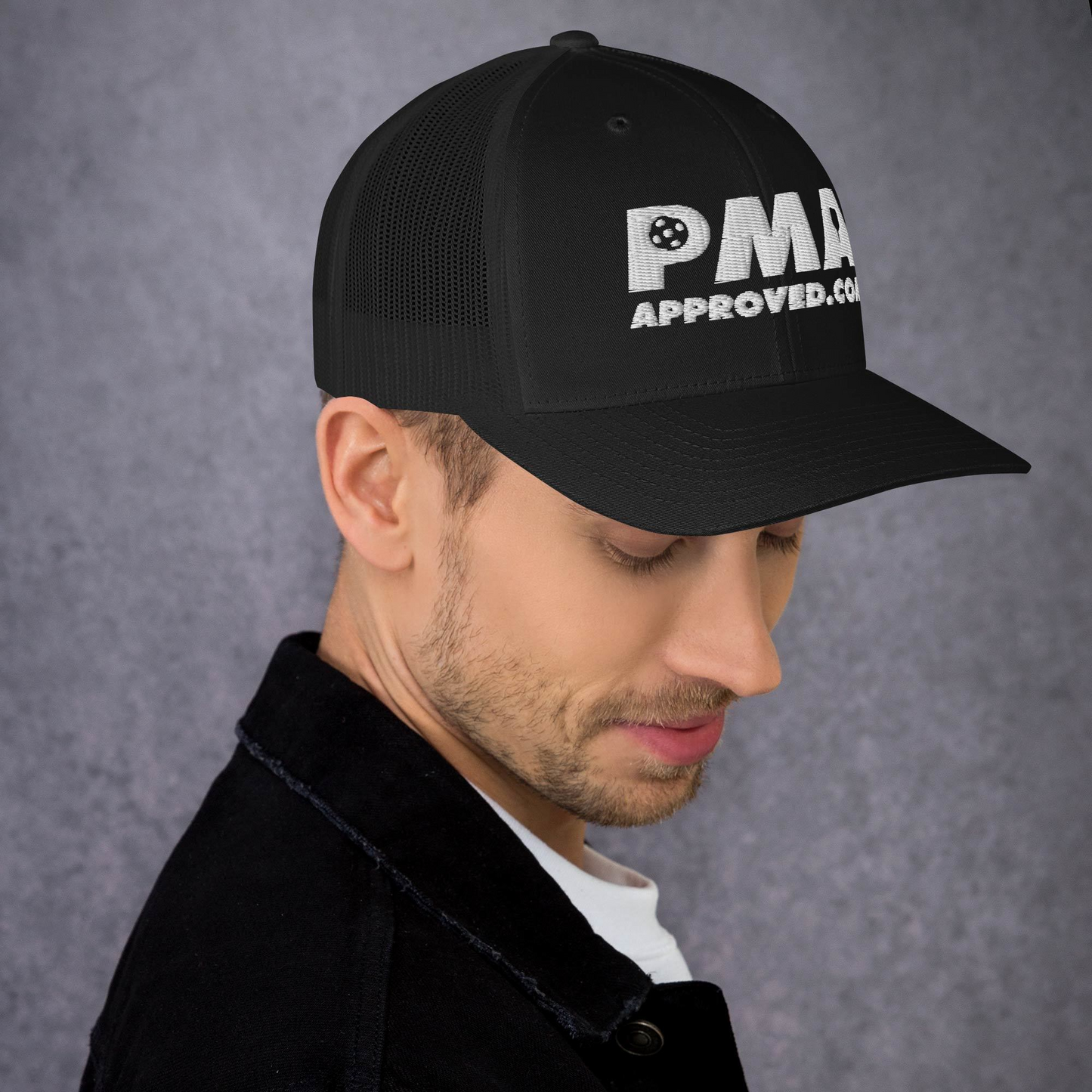 A man in a black Crown Pickleball Vintage Trucker Hat from the Pickleball Manufacturers Association, featuring “PMA APPROVED.COM” in white text, looks down with a slight smile against a textured gray background. The hat’s bill shades his face.