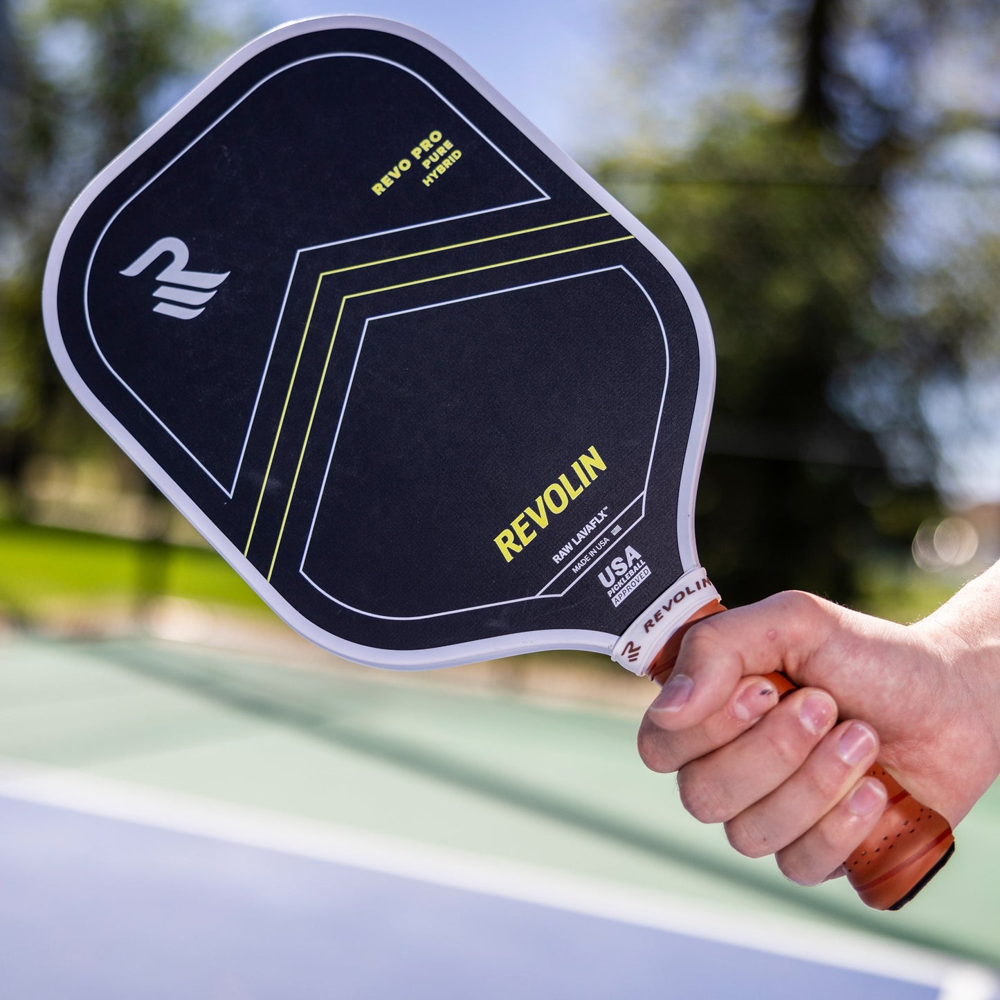 Someone holds a black Revolin Sports Revo Pro Pure Pickleball Paddle, made with basalt fiber and yellow accents, on an outdoor court. The paddle is angled close to the camera, with blurred trees and netting in the sunny background.