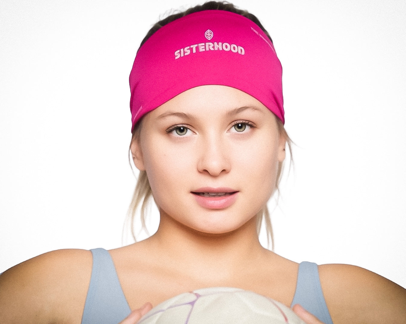A young woman with light skin and straight blond hair wears a pink, sweat-wicking Massov Skull Wrap. She has hazel eyes, a slight smile, and holds a white volleyball while dressed in a light blue tank top against a plain white background.