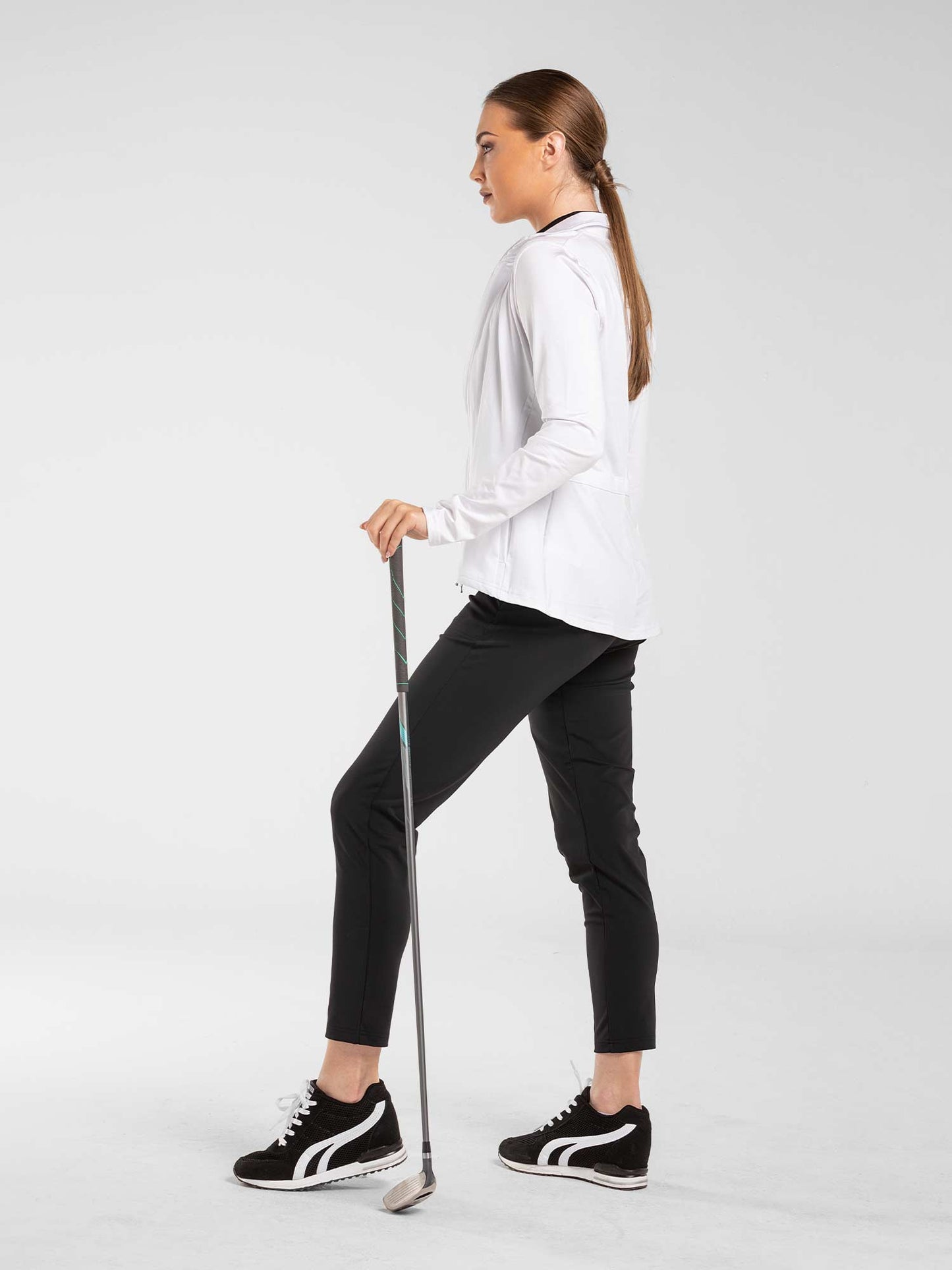 A woman stands in profile on a white background, holding a golf club. She wears a white long-sleeve top and inPhorm NYC's Stylish Women's Golf Pants—fitted and moisture-wicking—paired with black sneakers with white soles. Her hair is tied back.