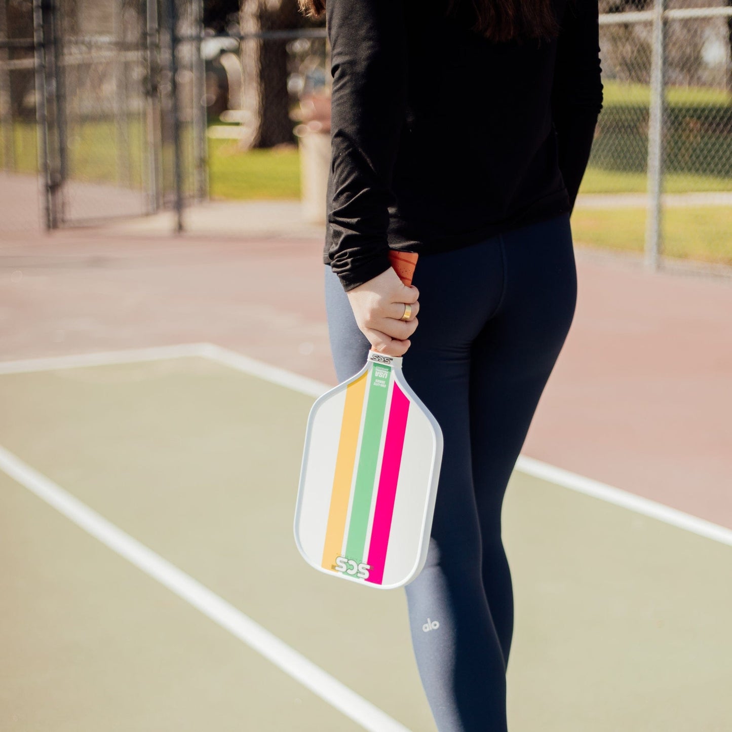 A person stands on a pickleball court, holding the Trifecta | Evo-Lite Series paddle by Salted City Sports. Shown from behind, only their lower half is visible; they're dressed in a black long-sleeve top and navy leggings.