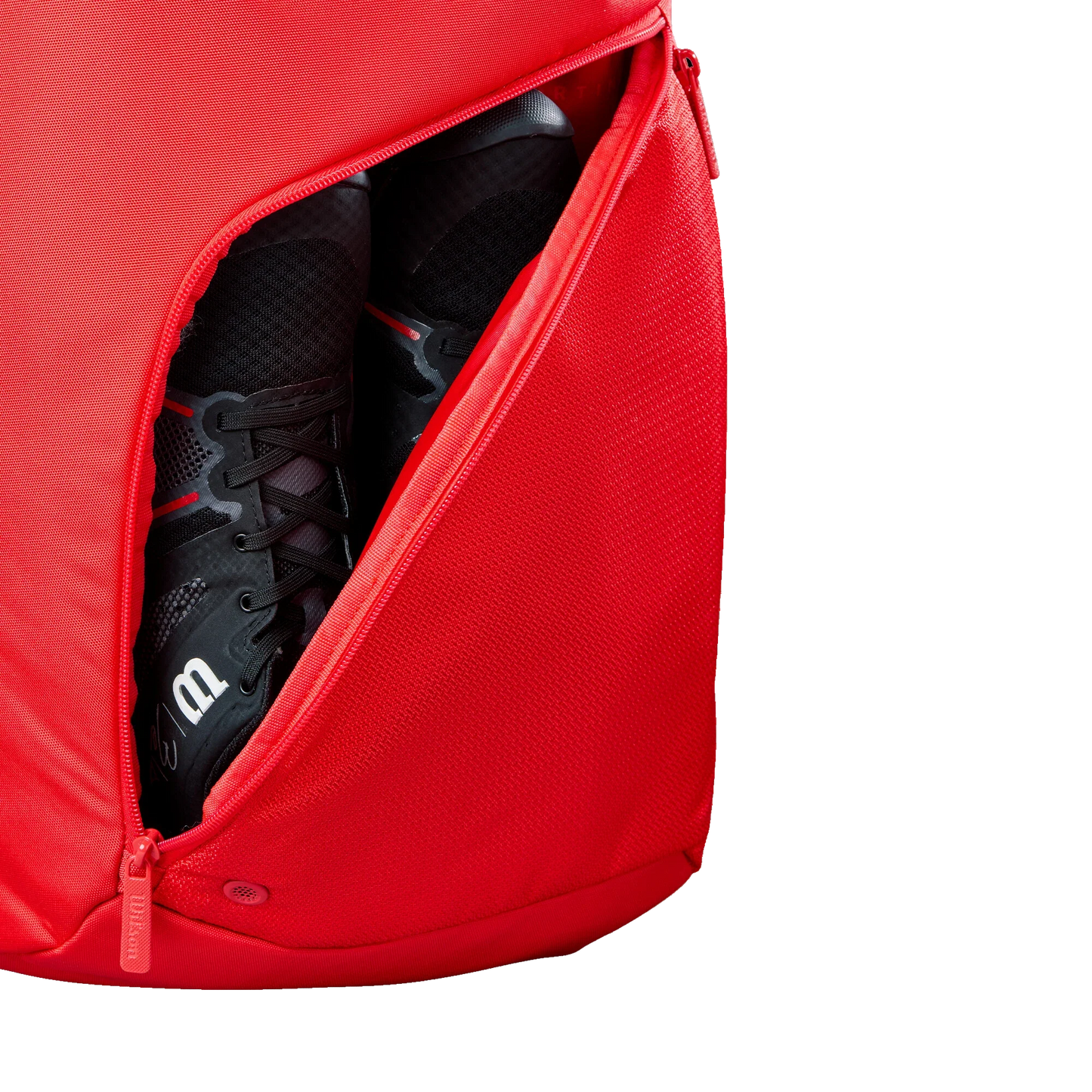 A close-up of the Wilson Tour Red Padel Backpack from Padel USA shows its diagonal zippered pocket partly open, revealing black athletic shoes inside. The plain white background highlights this stylish backpack and its contents.