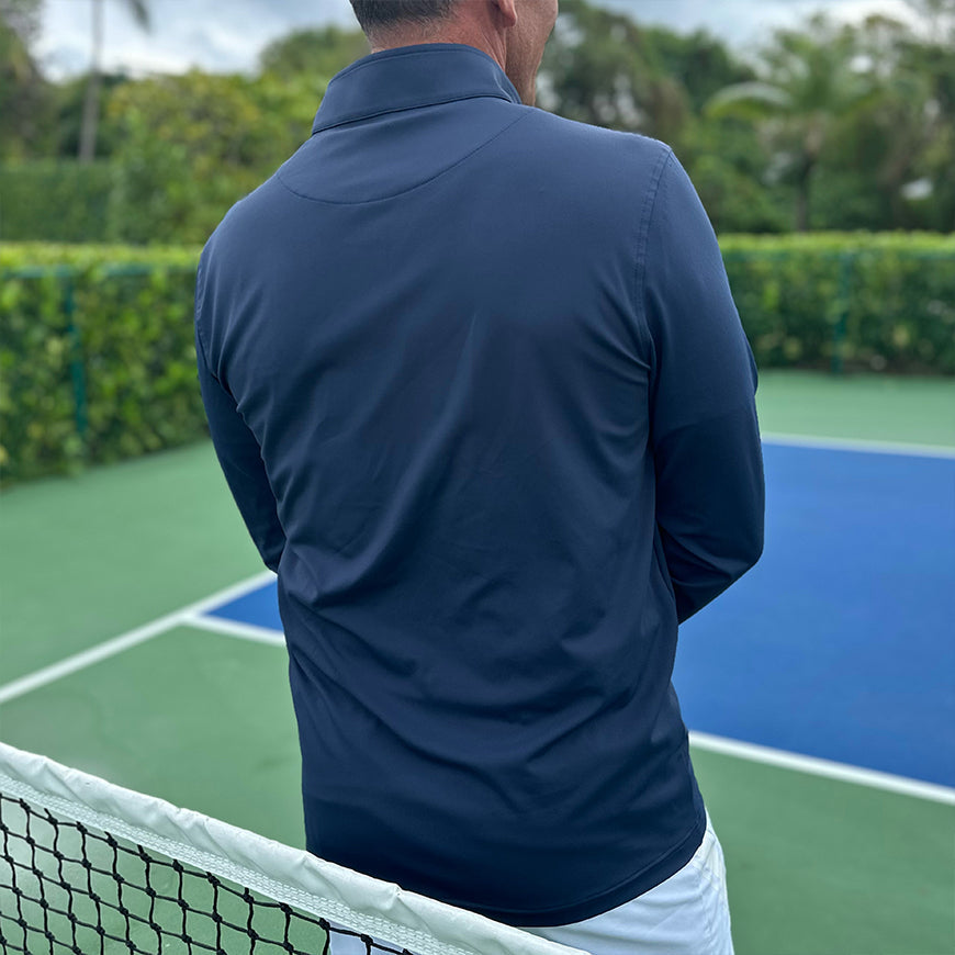 A man stands on a tennis court wearing the Swinton Pickleball Court Performance Quarter Zip Pullover in Navy and white shorts. Blue and green court lines and the net are visible, with lush trees bordering the scene.