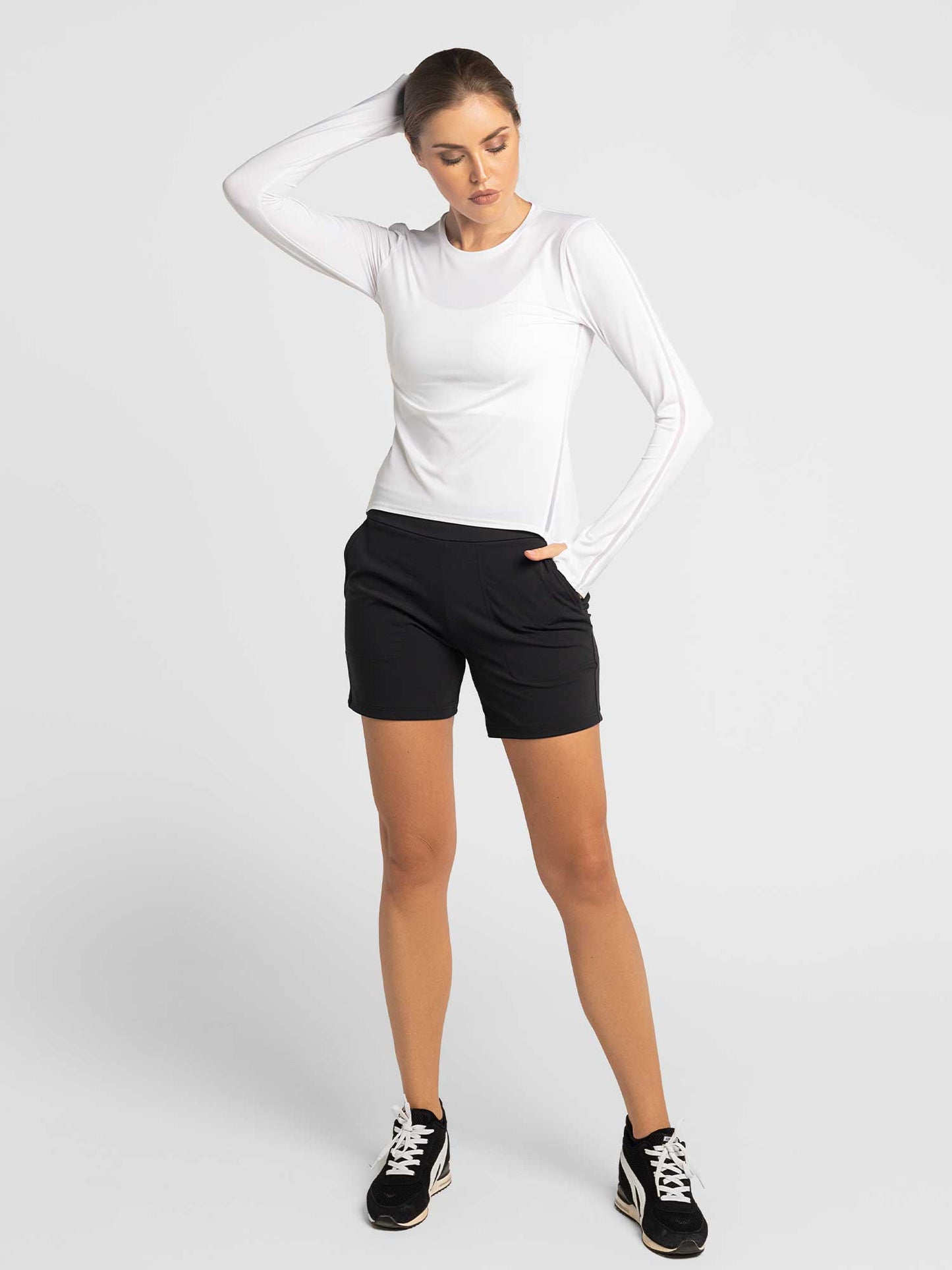 A woman poses against a white background in a fitted long-sleeve white top, black sneakers, and the inPhorm NYC Women's Cleo Short—black, stylish, and functional for active lifestyles—with one hand in her pocket and a neutral expression.