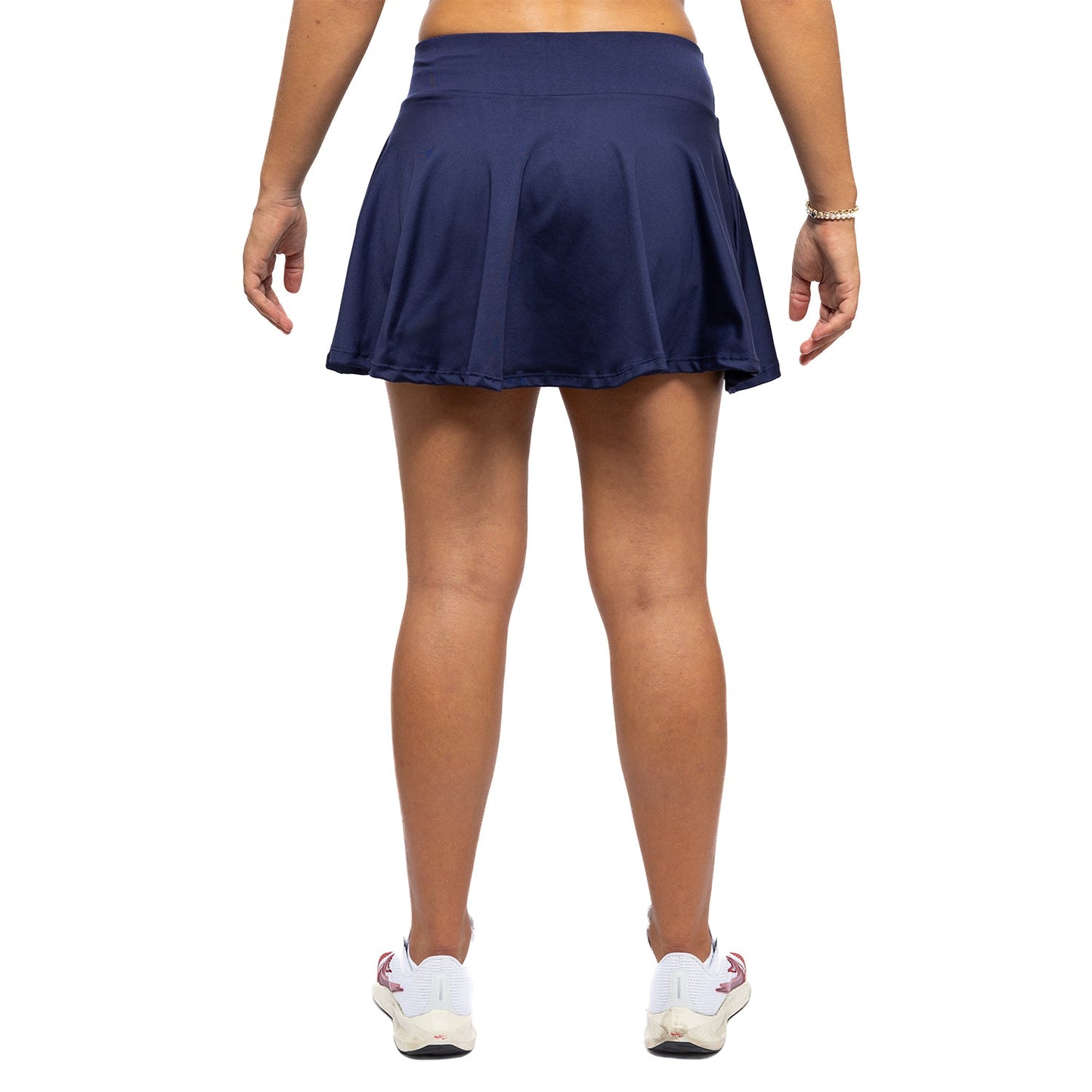 A model showcases the Diadem Sports Women's Essential Club Skirt in navy blue, paired with white sneakers with pink accents. Only the lower body is visible against a plain white background; a thin bracelet adorns the left wrist.