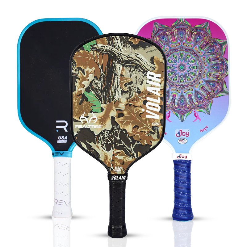 Three pickleball paddles stand upright. The left has a black face with blue trim, the middle has a camouflage pattern, and the right features a colorful mandala design with pink, blue, and purple accents.