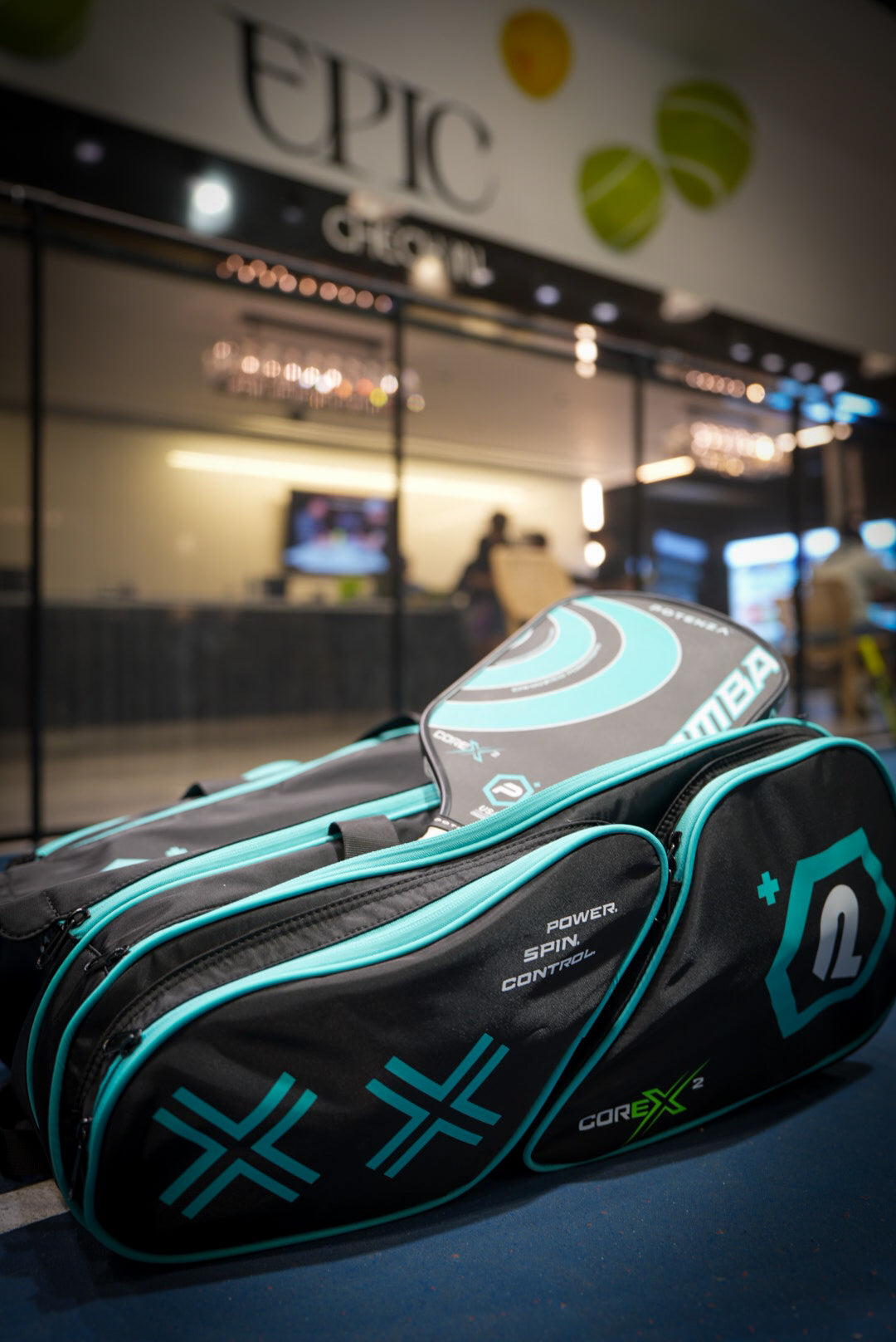 A black and turquoise Potenza Pickleball Tournament Paddle Bag and padel racket sit on the court floor before glass doors. The bag reads: POWER. SPIN. CONTROL. Behind them, a modern lounge glows with an illuminated EPIC sign overhead.
