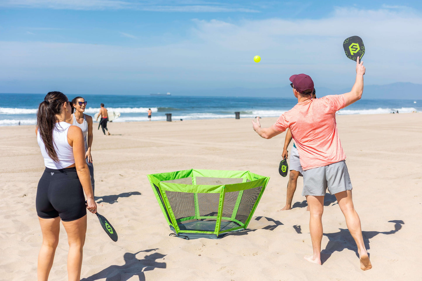 Four people play PaddleSmash by PaddleSmash on a sandy beach near the ocean. They stand around the green net, each holding a paddle as the yellow ball flies through blue skies, with others enjoying the game in the background.