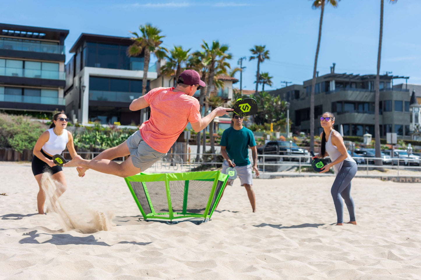 Four friends play PaddleSmash by the sea, leaping and hitting volleys over a green net near beach houses and palms. Sand flies as they enjoy this energetic outdoor game from PaddleSmash, delivering backyard Pickleball fun on the coast.