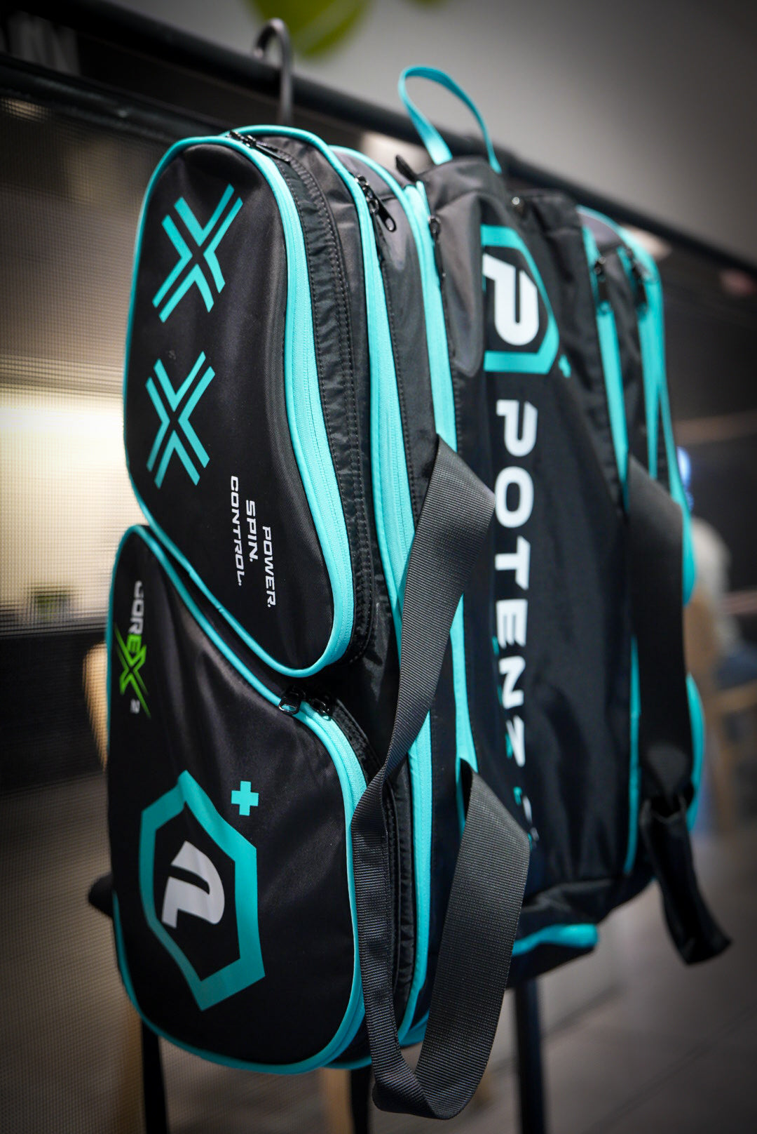 The Potenza Pickleball Tournament Paddle Bag, in black with teal and white geometric designs, hangs on an indoor railing. This bag features multiple compartments, visible zippers, paddle protection, and bold brand logos.