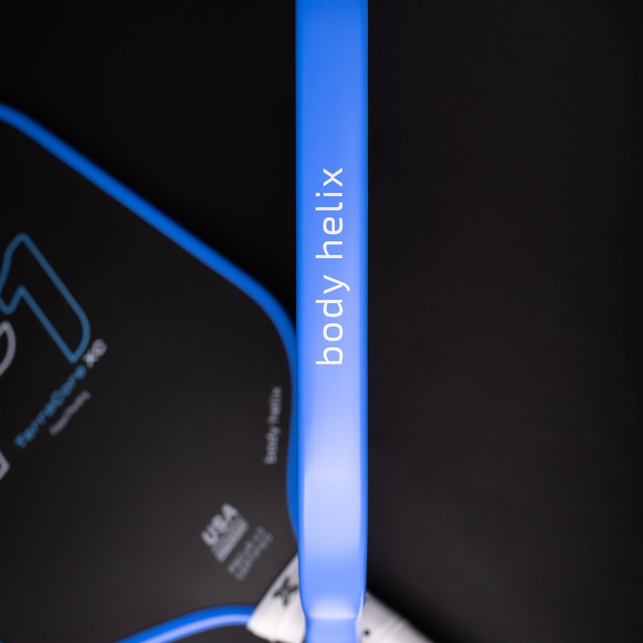 Close-up of the blue FLiK F1 Elongated Pickleball Paddle by body helix, showing its edge with the brand name in vertical print. The mostly black background reveals part of the USAP-approved paddle’s face with white text and curved blue trim.