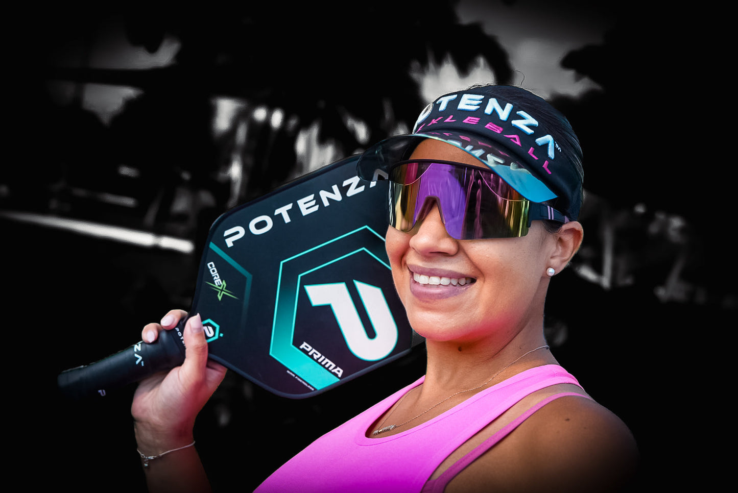 A smiling woman in a pink tank top, visor, and reflective sunglasses holds a Potenza Pickleball PRIMA COREx paddle over her shoulder. The black-and-white background highlights her and the colorful PRIMA COREx paddle.