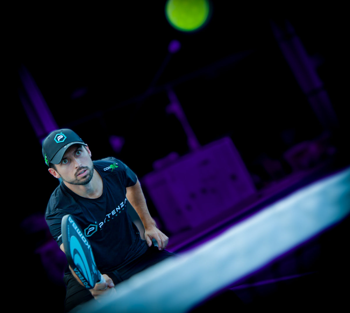 A focused man in a black hat and shirt prepares to hit a pickleball on court with the Potenza Pickleball KOMBA+ COREx2 carbon fiber paddle. A neon green ball and purple-blue lighting enhance the action and his intensity.