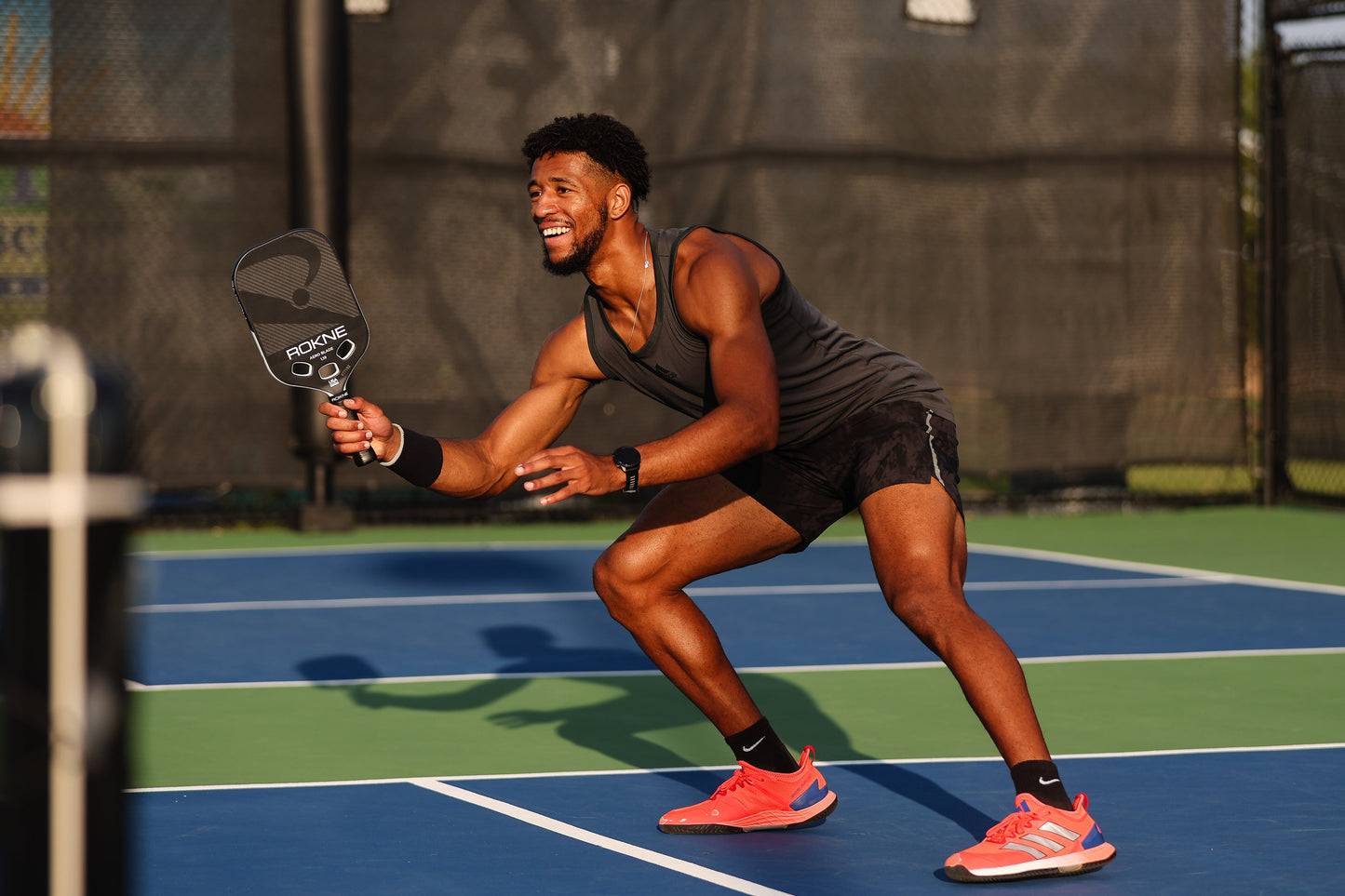 A man in athletic wear smiles while playing pickleball on an outdoor court, crouched and ready with his ROKNE AERO BLADE 1.19 paddle. Sunlight highlights his dark tank top, black shorts, and bright orange sneakers on the blue-green court.