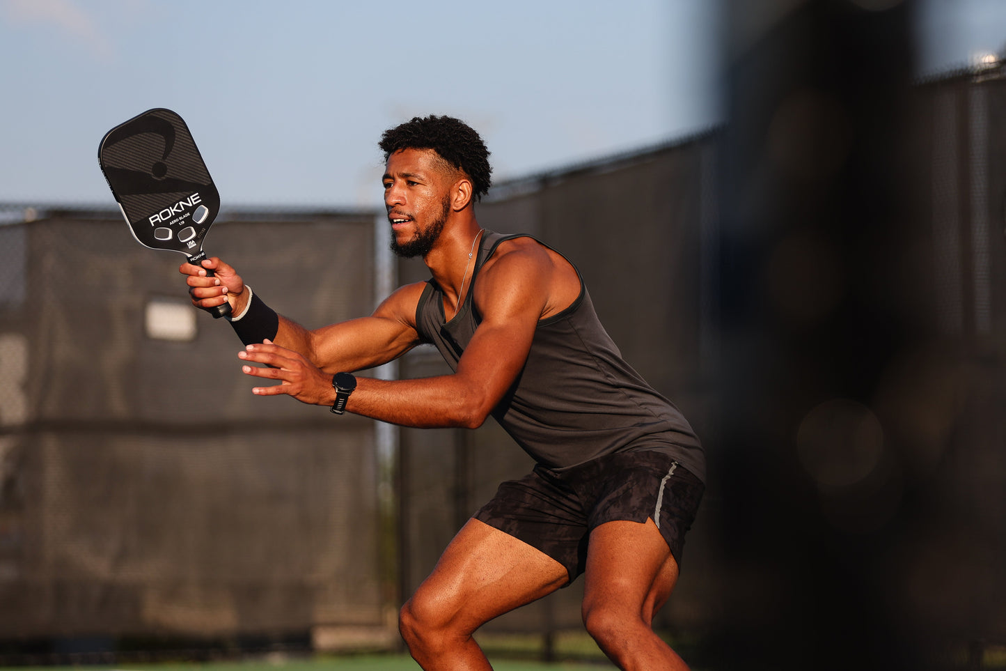 A man in a sleeveless dark athletic shirt and shorts plays pickleball outdoors, gripping a ROKNE AERO BLADE 1.19 paddle with carbon fiber mono-frame in his right hand, knees bent and focused as he prepares to return a shot under the clear sky.