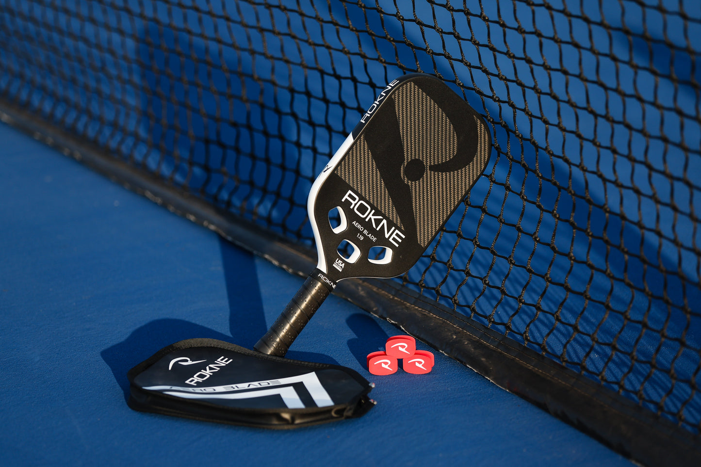 A black ROKNE AERO BLADE 1.19 paddle with a carbon fiber mono-frame leans against a net on a blue court, beside a matching cover and two red pickleball tokens, all illuminated by the late afternoon sun.