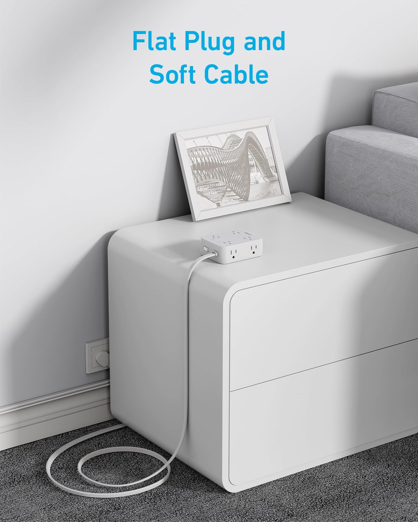 The Anker 341 USB Power Strip, featuring surge protection, a flat plug, and a soft flexible cable, sits on a modern nightstand with its cable neatly looped on gray carpet. A black-and-white photo leans above. Blue text: Flat Plug and Soft Cable.