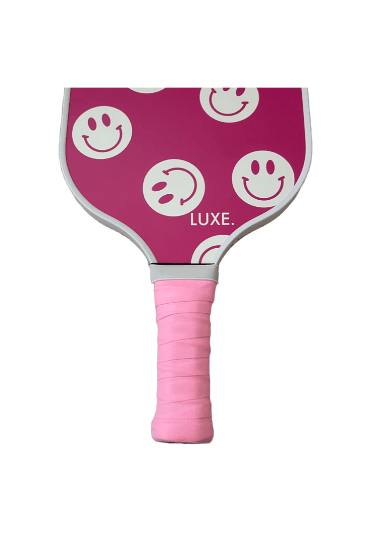 A pink Pickleball Paddle Grip by LUXE Pickleball features a light pink overgrip, white smiley faces (one winking) outlined in pink on the paddle face, and "LUXE." in white near the bottom.