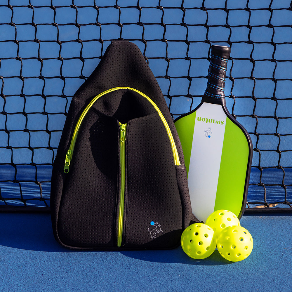 The Swinton Pickleball Starter Bundle - Black Sling Bag Combo rests on a blue court by the net, beside a green and white paddle and three yellow pickleballs—the perfect pick for stylish pickleball accessories.