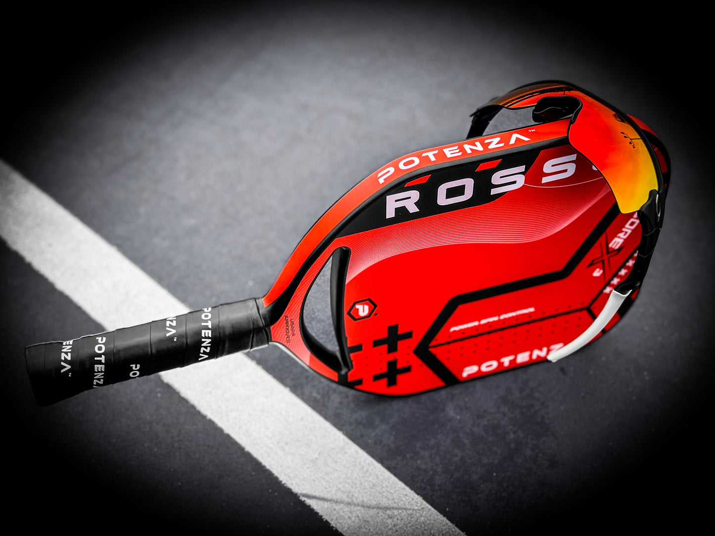 A ROSSO+ COREx3 pickleball paddle from Potenza Pickleball, featuring red and black colors with COREx3 tech and a black grip, lies on a dark court near the white boundary. Red-orange sports sunglasses rest on the professional paddle.