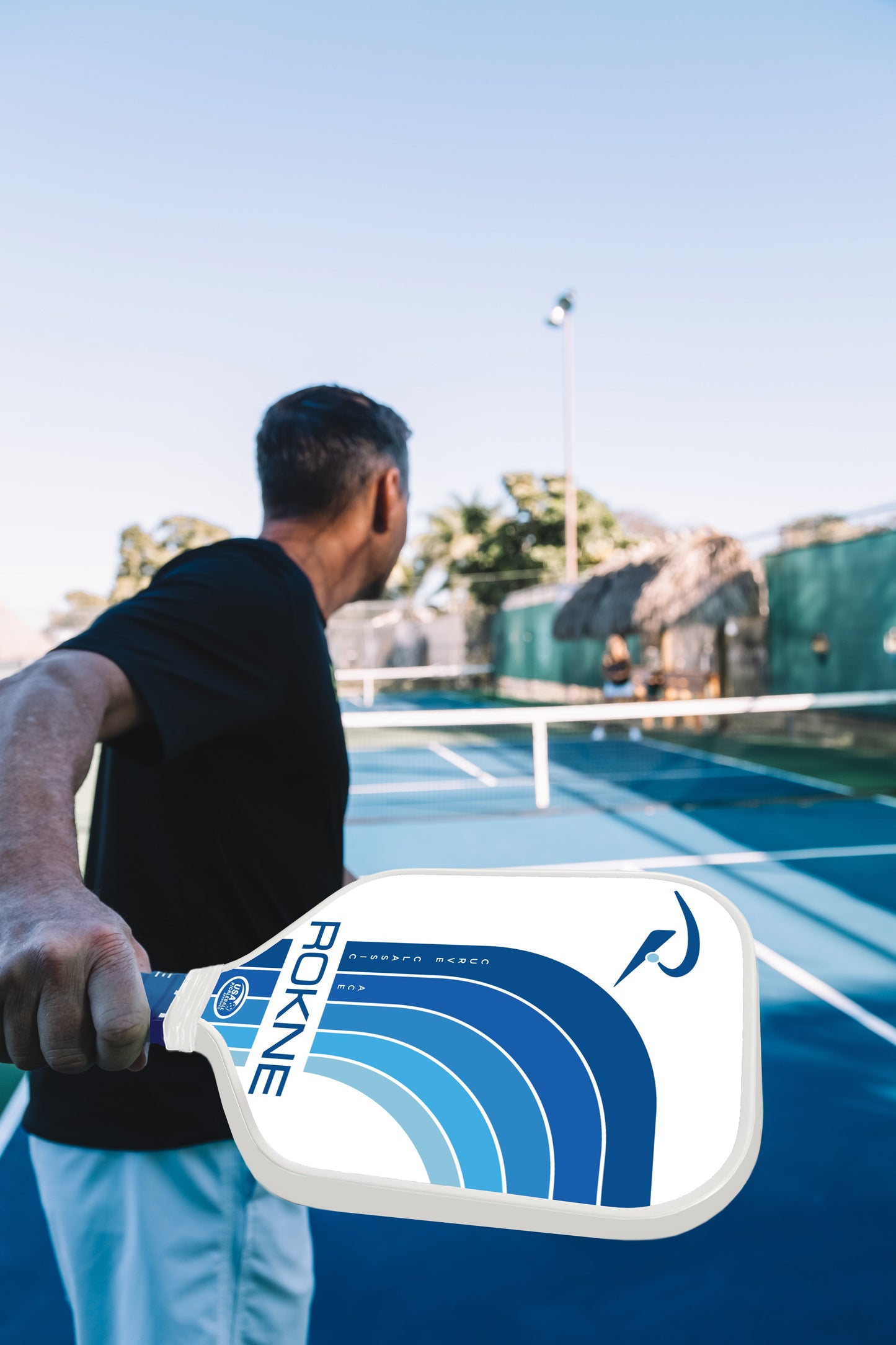 A man in a black shirt gets ready to hit a pickleball with the ROKNE CURVE CLASSIC ACE paddle, which has a textured fiberglass surface, on an outdoor court. Green fencing, trees, and another player are visible under a clear sky.