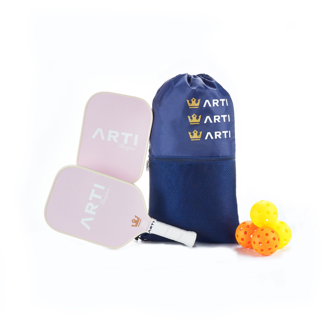 The ARTI Fiberglass "Valentine’s" Pickleball Paddle Set features two light pink paddles with white handles, a blue ARTI drawstring bag with gold crowns, plus three yellow and one orange perforated balls on a white background.
