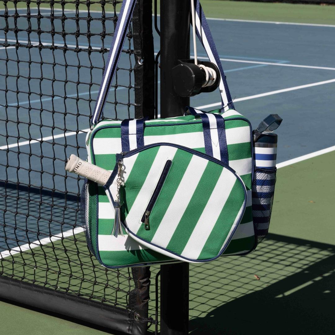The Fresh Pickle Lola Pickleball Tote Bag in green and white stripes with navy accents hangs on a blue and green court net post, holding a racket in a zip pocket and a striped water bottle—ideal for carrying all your pickleball essentials.