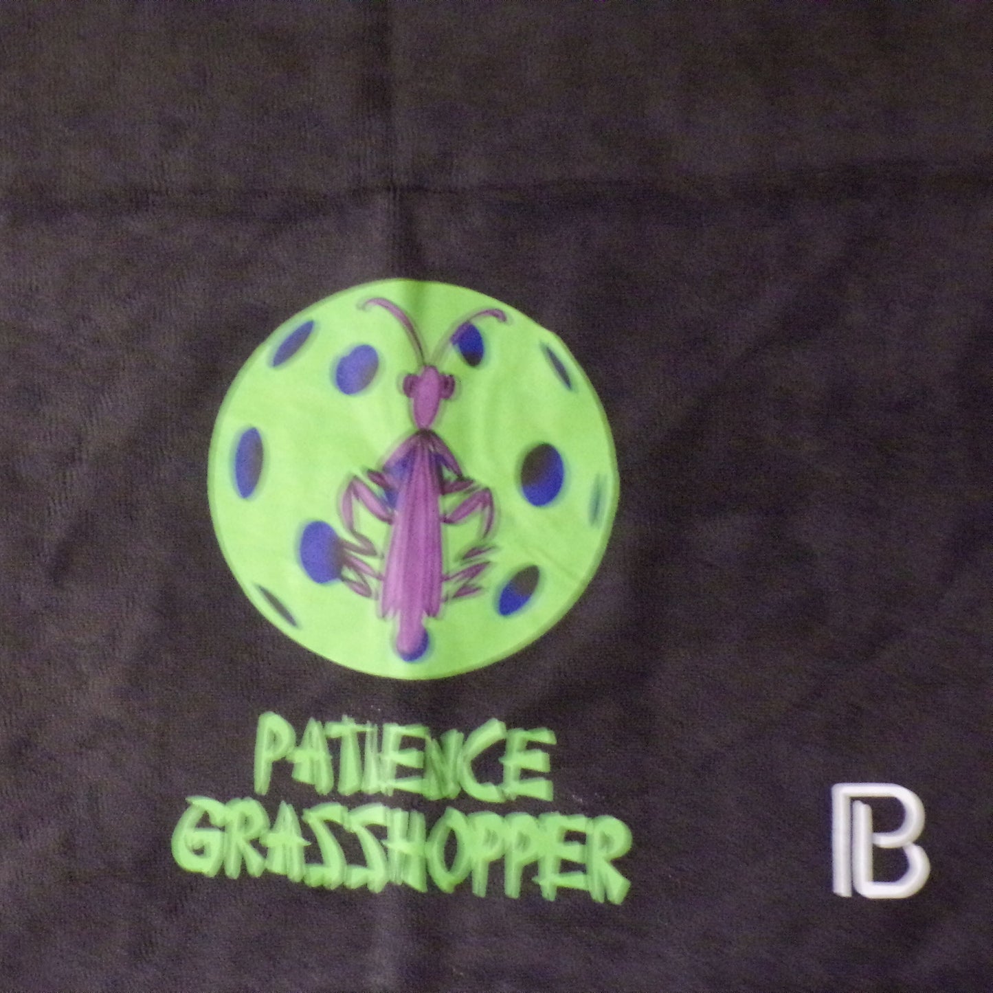A green circle with blue spots and a purple grasshopper illustration sits above green text reading PATIENCE GRASSHOPPER. A white B logo is in the corner, all on black polyester fabric. This is the Pickleball Bella Patience Pickleball Towel by Pickleball Bella.