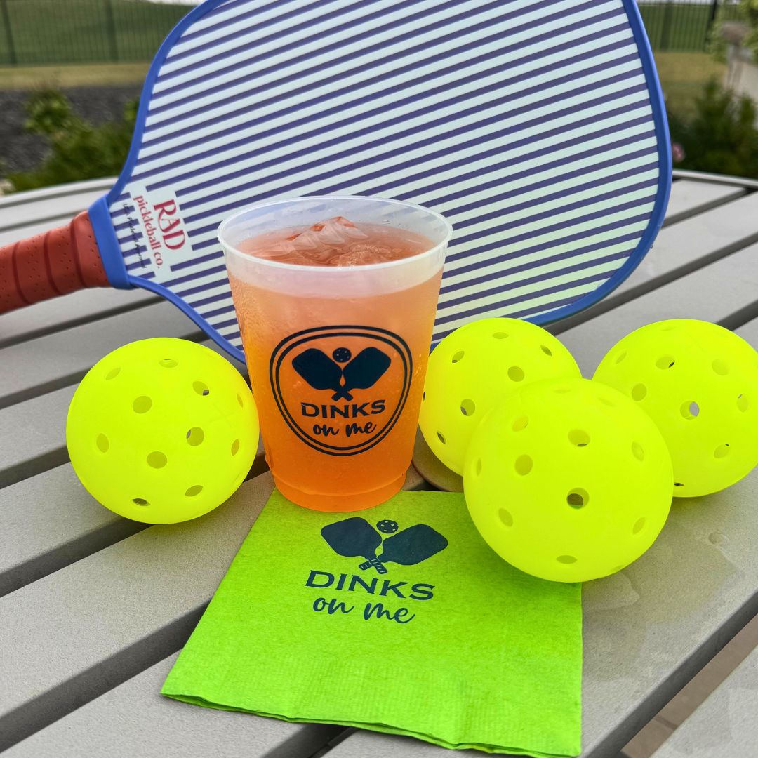 A shatterproof Fresh Pickle Pickleball Roadie Cup with orange drink and ice rests on a green "DINKS on me" napkin, surrounded by yellow pickleballs and a blue striped paddle on an outdoor gray table.