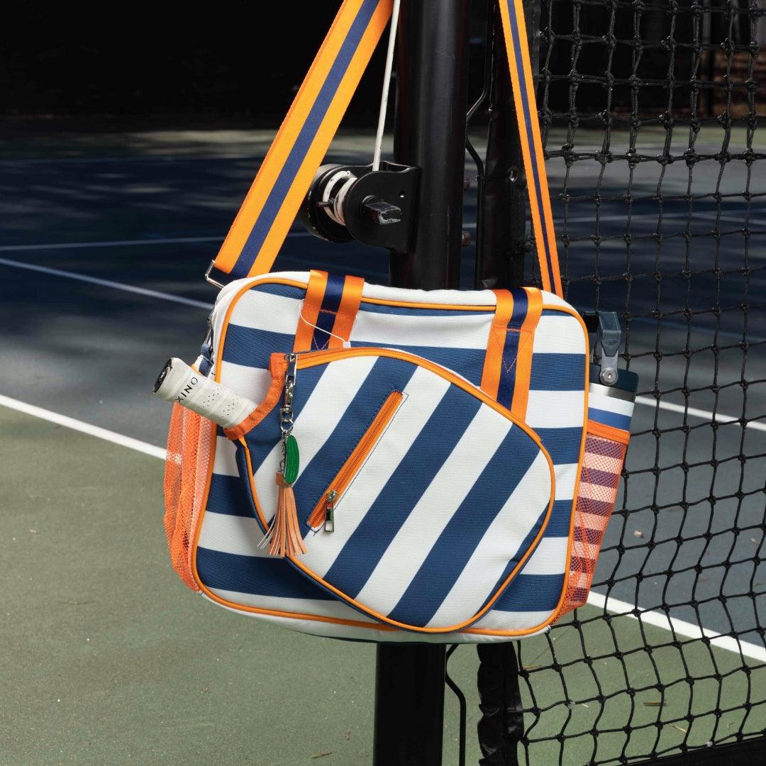 The Fresh Pickle Lola Pickleball Tote Bag in blue and white stripes with orange accents hangs on a black net post. It features a front pocket, orange tassel zipper, green keychain, and side pocket for holding accessories.
