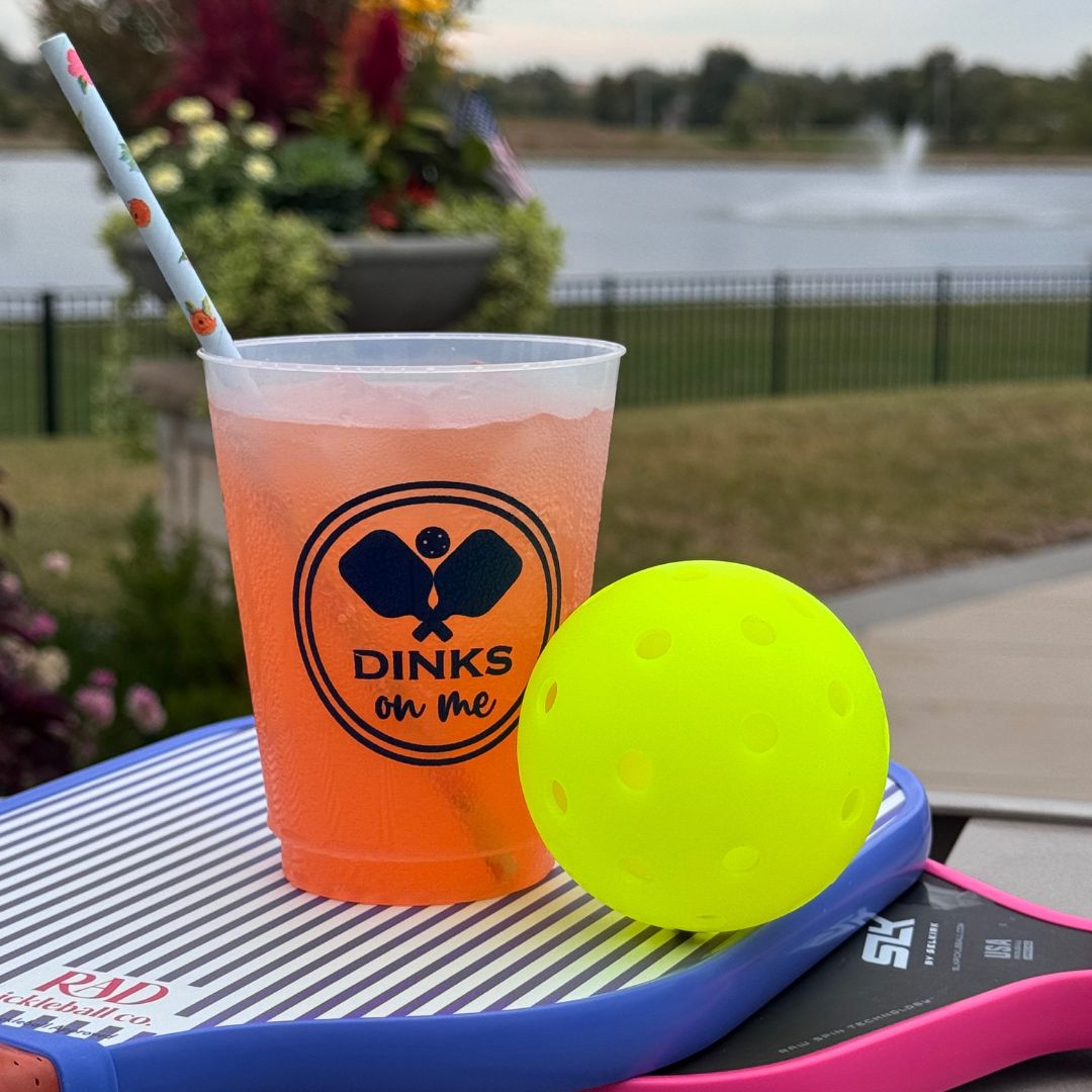 A Fresh Pickle Pickleball Roadie Cup with a reddish-orange drink and striped straw sits on two paddles, labeled “DINKS on ME.” A yellow pickleball is nearby, with a lake, fountain, fence, and greenery in the background.