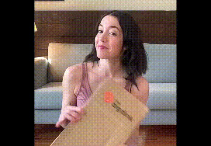 A woman with shoulder-length dark hair, wearing a sleeveless pink top, sits on a couch and eagerly opens a large brown envelope, surprised and amused to find it empty—no The Big Dink Bundle carbon fiber pickleball paddles inside!.