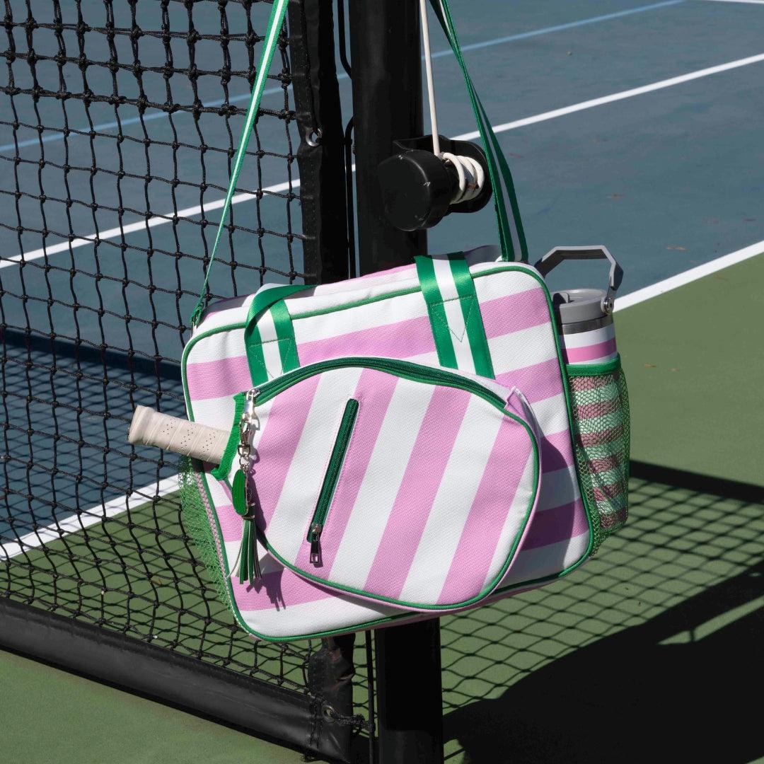 The Fresh Pickle Lola Pickleball Tote Bag, pink and white striped with green accents, hangs on a black tennis net post. It holds a racquet and silver water bottle, with a blue and green tennis court in the background.