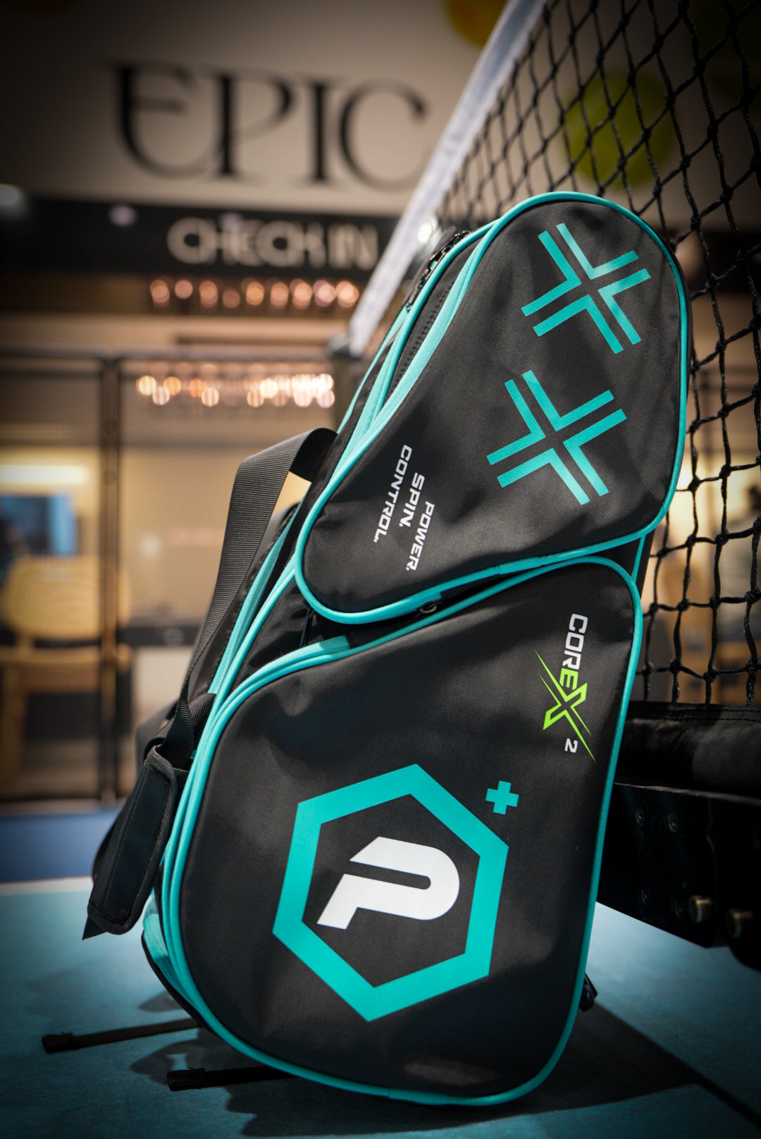 A black and teal Potenza Pickleball Tournament Paddle Bag labeled CORE X and P rests against an indoor net, showcasing bold graphics, zippers, paddle protection compartments, and a blurred EPIC CHECK IN sign in the background.